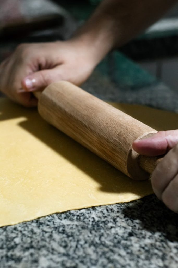 Close-up of hands using a rolling pin to flatten homemade pasta dough indoors.